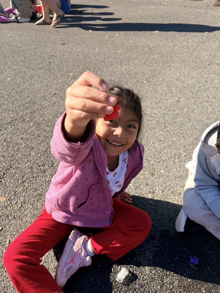 A smiling student in a pink jacket sits on the playground and holds up a small red object she uncovered during the activity.