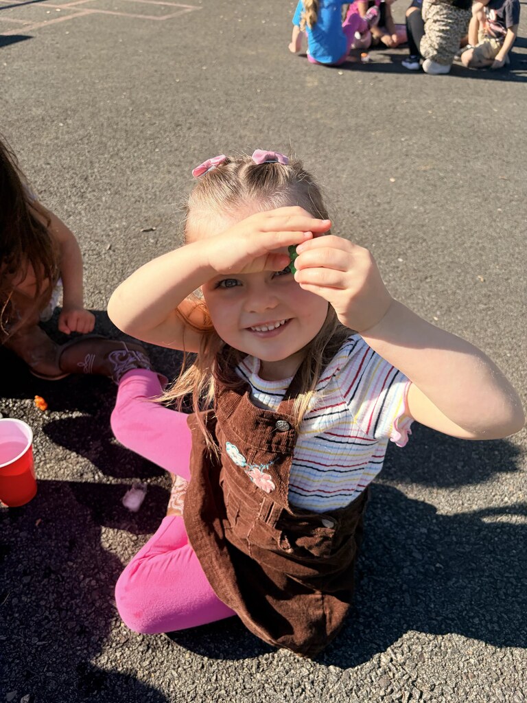 A smiling student sits on the playground and proudly holds up a small green object created during the activity.