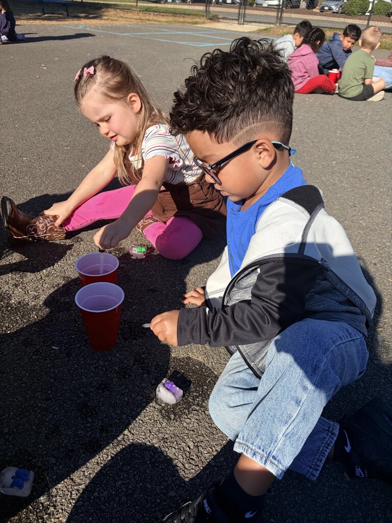 Two students sit on the playground using droppers and cups of water during a hands-on activity while other classmates work nearby.