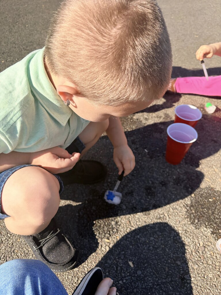 A student crouches on the playground and uses a dropper to add liquid to a small blue object resting on a piece of ice.