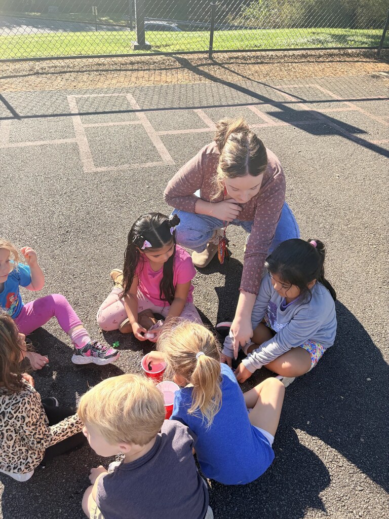 Students and a teacher sit together on the playground pavement while participating in a hands-on activity using cups of water, droppers, ice, and small colorful objects.