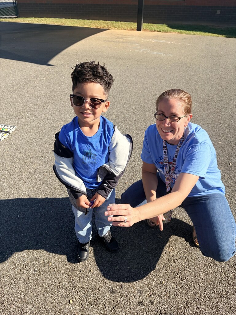 A smiling student wearing sunglasses stands beside a teacher who holds up a small container during an outdoor classroom activity.