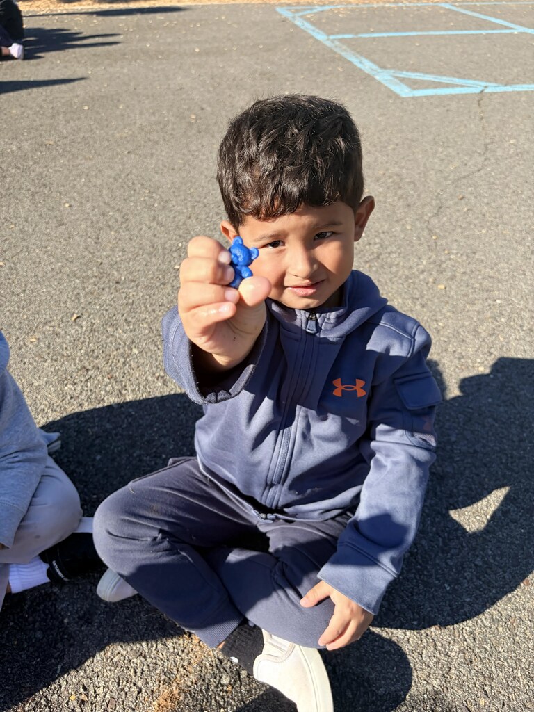 A smiling student sits near the playground equipment and proudly displays a small red object while holding a dropper in his other hand.