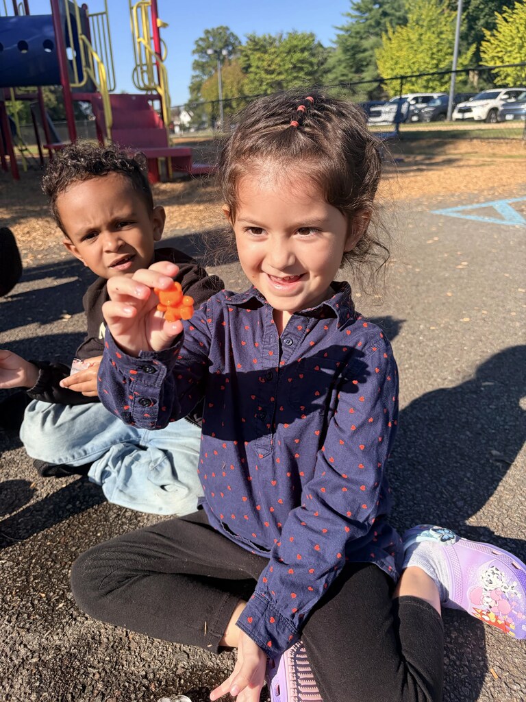 A smiling student sits on the playground and holds up a small orange object she uncovered during the hands-on activity.