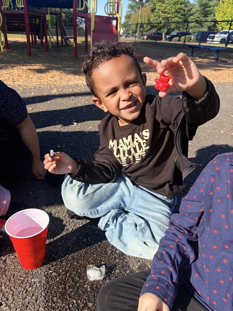 A smiling student sits near the playground equipment and proudly displays a small red object while holding a dropper in his other hand.