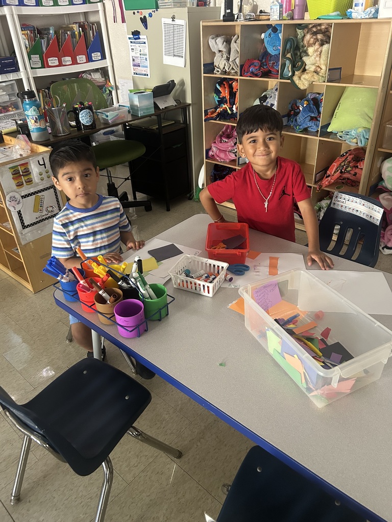 Two children sit at a table in a classroom, surrounded by art supplies and materials.