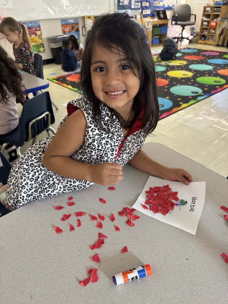 A young girl sits at a table, smiling while working with a paper and red petals.