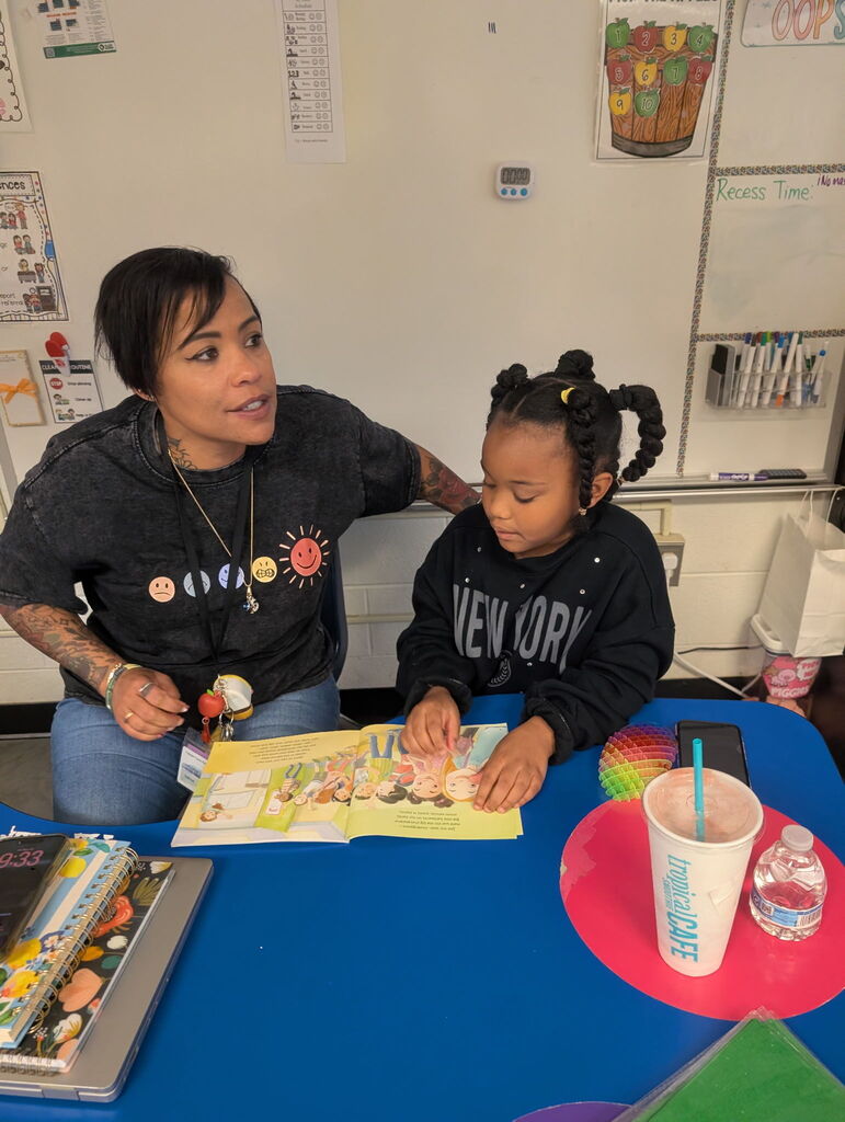 Woman and child seated at a table, reading a book. Table has a cup, bottle, and books.