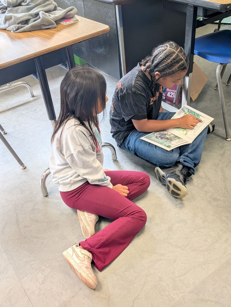 Two children sit on the floor, one reading a book. The girl wears white shirt, red pants, and sneakers. The other wears a black shirt, blue jeans, and boots.