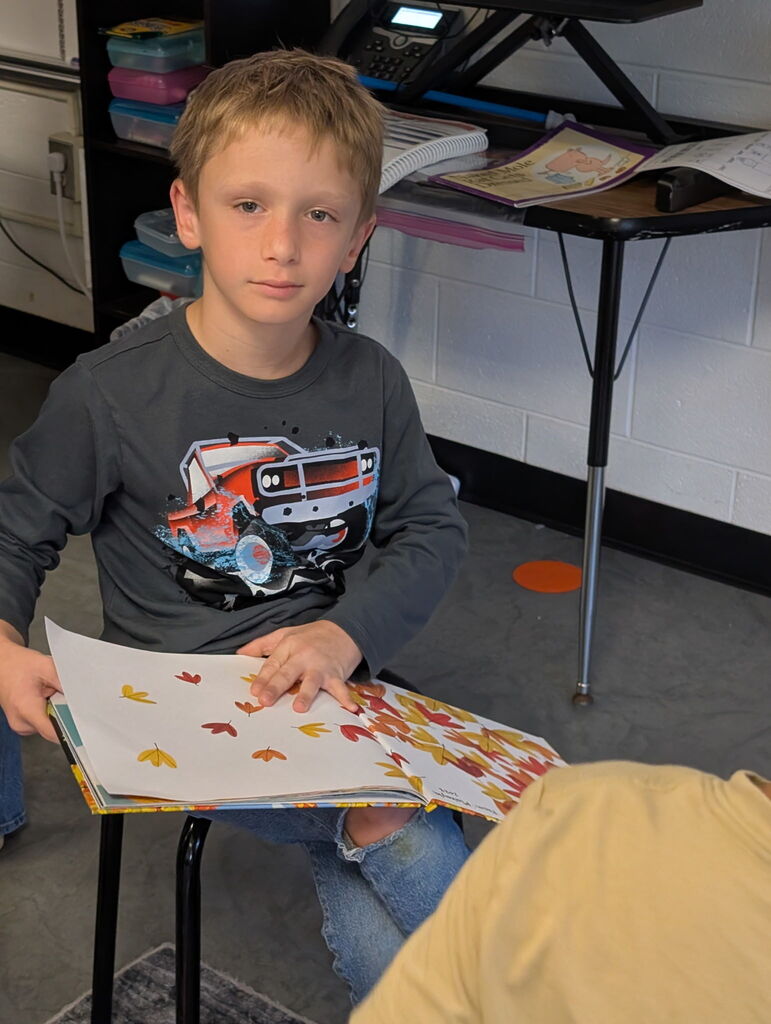 A boy in a black shirt reads a book in a classroom. The book has a red truck image.