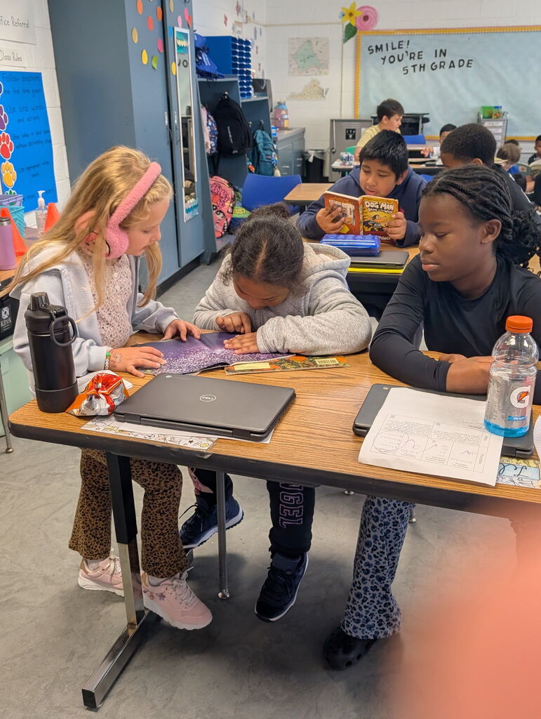 Three children at a desk in a classroom. One child is reading a book. Another is working on a laptop.