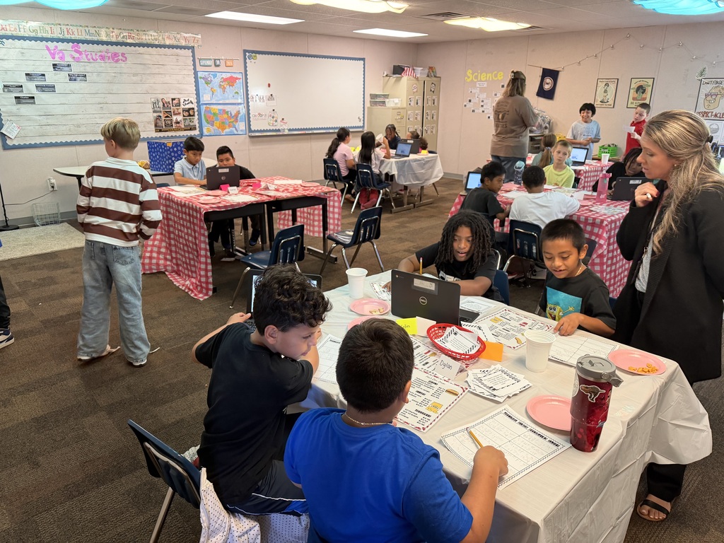 Several children sit at tables with laptops and papers. A woman stands nearby and speaks to the group.