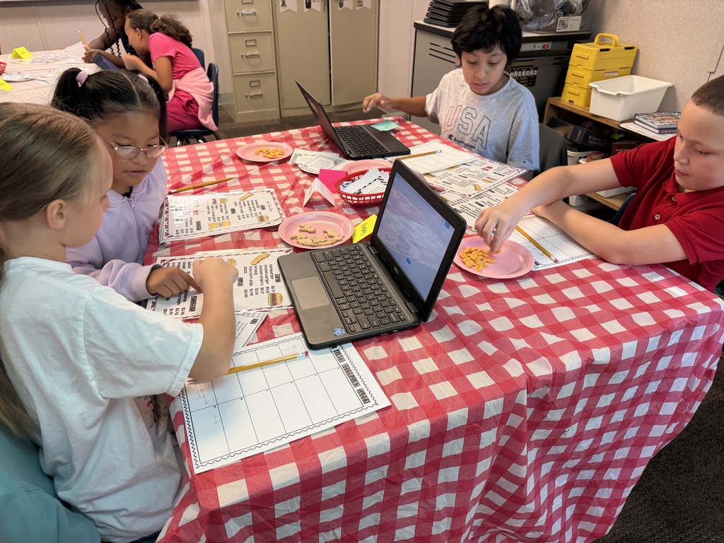Four children in a classroom are seated at a table with laptops. They are looking at a screen.
