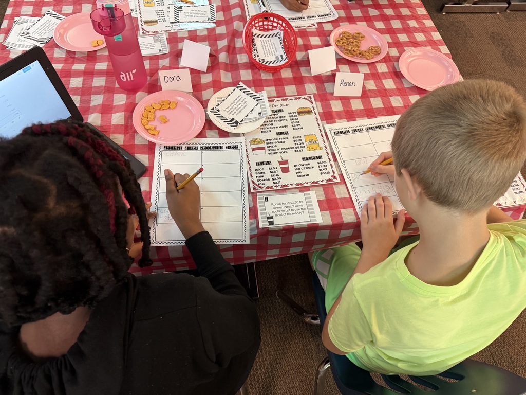 Two children at a table, one with braided hair, the other in a yellow shirt, both writing. A laptop, plates, and a water bottle are on the table.