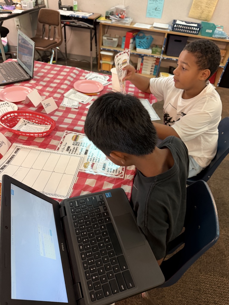 Two boys in a classroom, seated at a red-and-white checkered table with laptops, one holding cards.