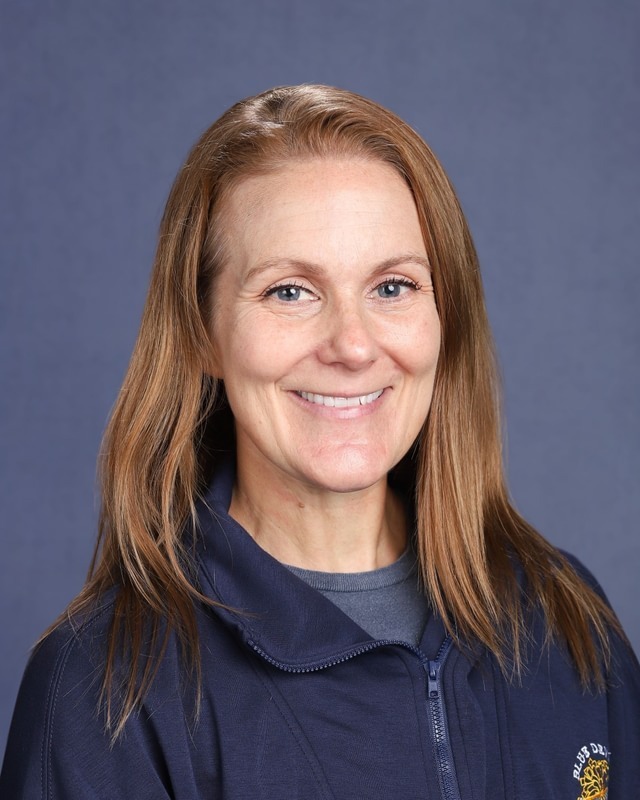 A smiling woman with long blonde hair in a navy blue jacket. The background is a plain blue wall.