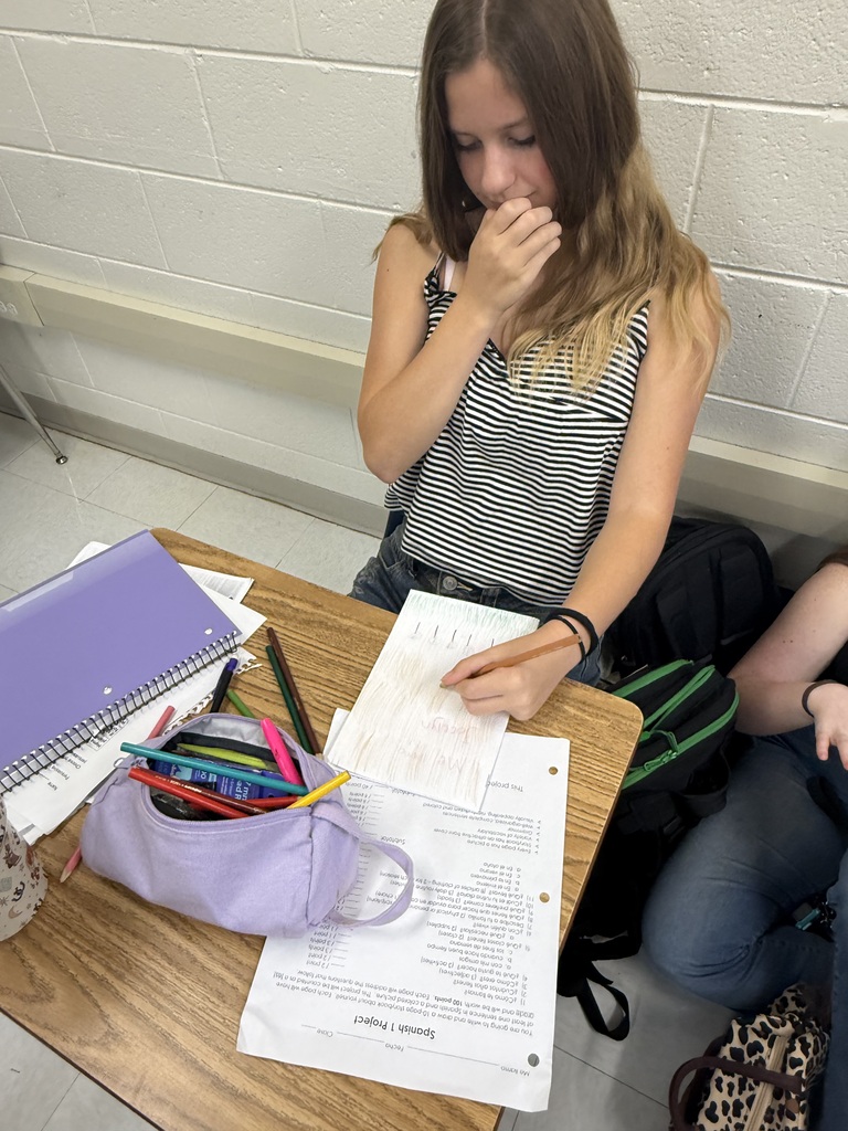 A person sits at a table, covering their mouth, with a notebook, pens, and a bag.