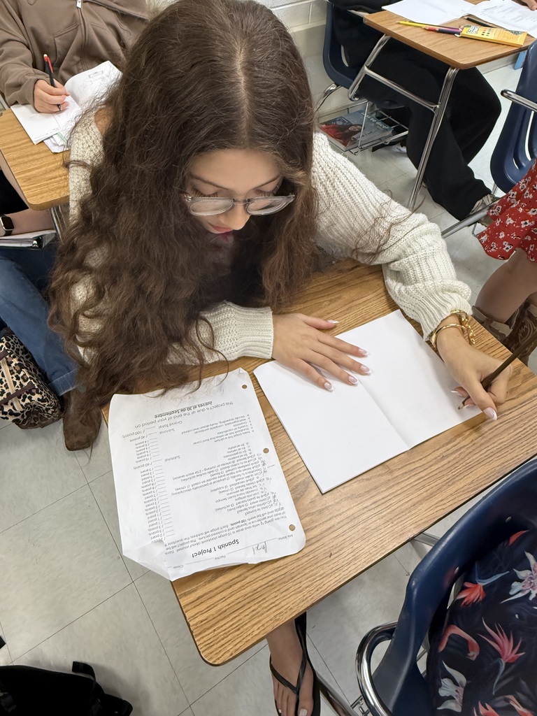 Person with glasses and long hair writing in a classroom with papers and books on desk.