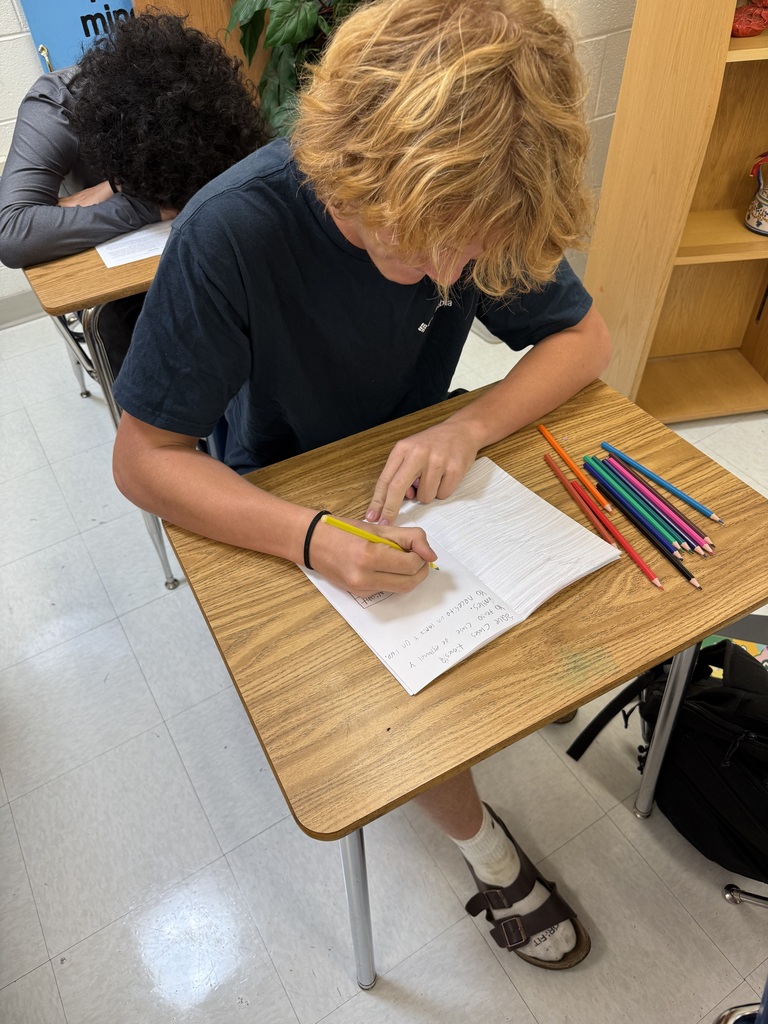A student seated at a desk, writing on a sheet of paper with a pencil. Next to him, a bag and a shelf.