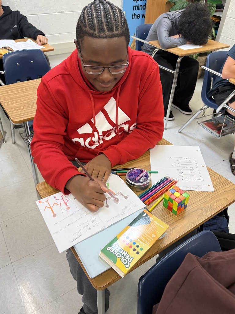 Person in red hoodie seated at desk writing on paper, surrounded by colored pencils and a Rubik's Cube.