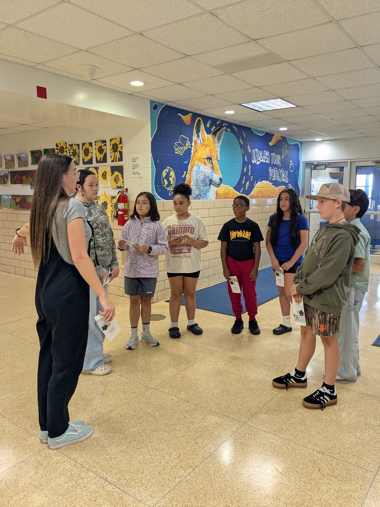 A group of students, one holding papers, stands in a hallway. A mural on the wall features a fox.