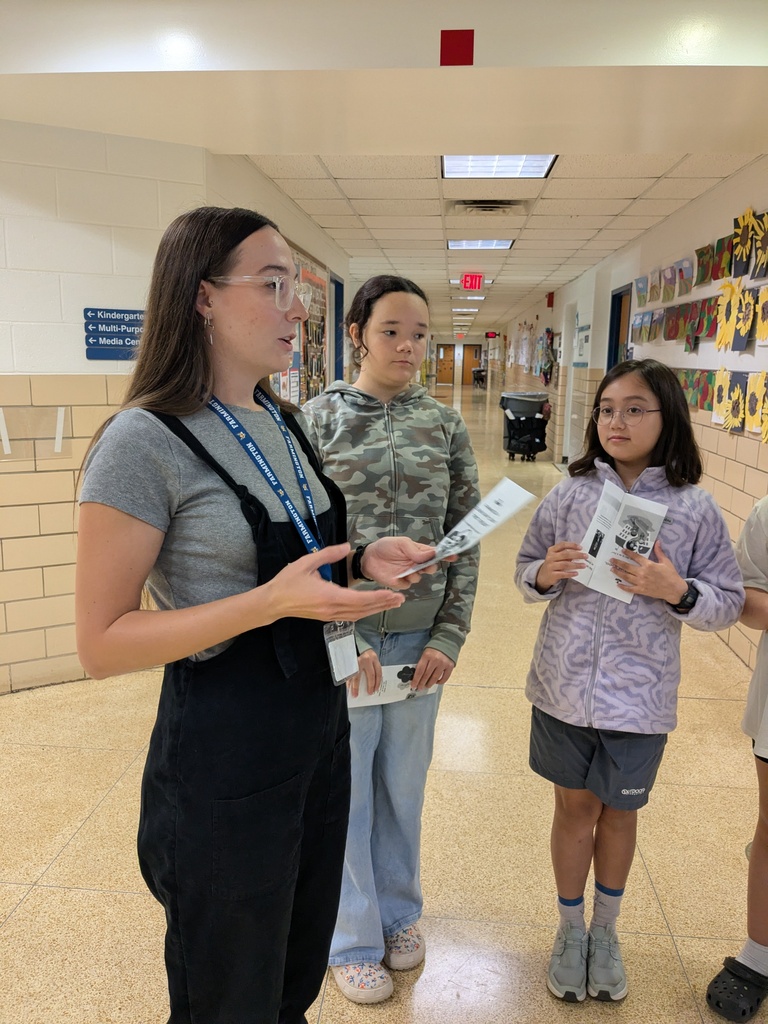 Three people in a hallway, one holding a paper and speaking, another holding a book, and a third holding a magazine.