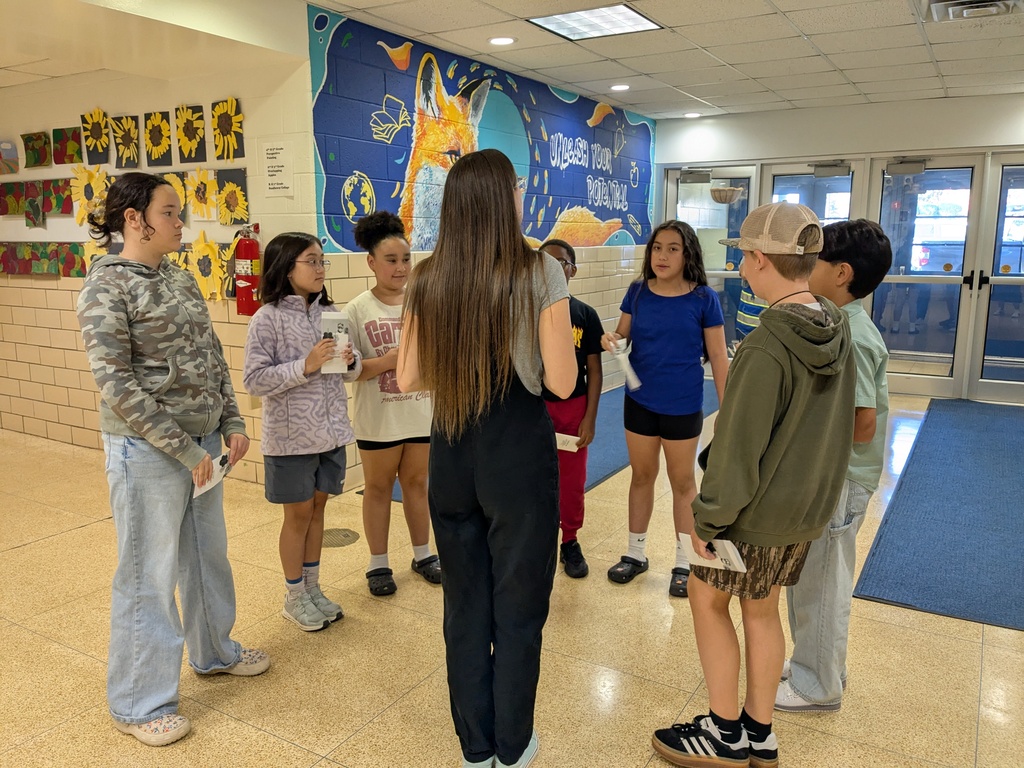 Several children in a school hallway, two holding papers, standing in a circle under a mural.