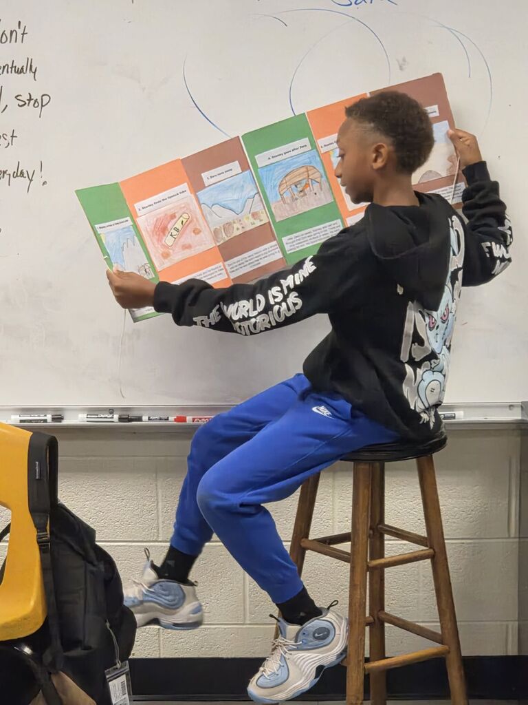 A student in blue pants and a black sweatshirt holds a book up to a whiteboard.