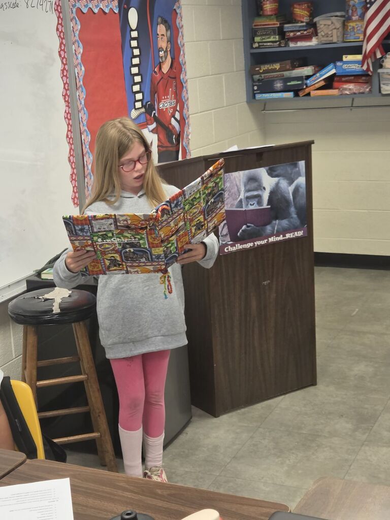 A young girl with glasses holds an open comic book while standing in a classroom. Behind her, a podium and shelves are visible.
