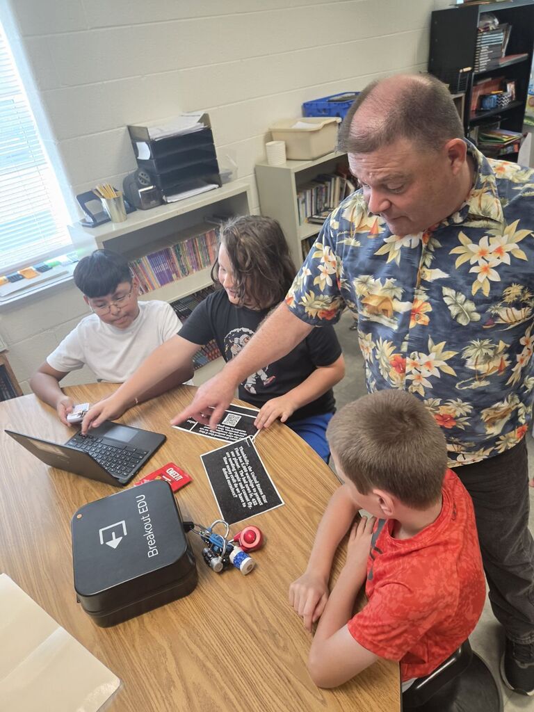 A man shows a laptop and other items on a table to two children. The setting is a classroom.