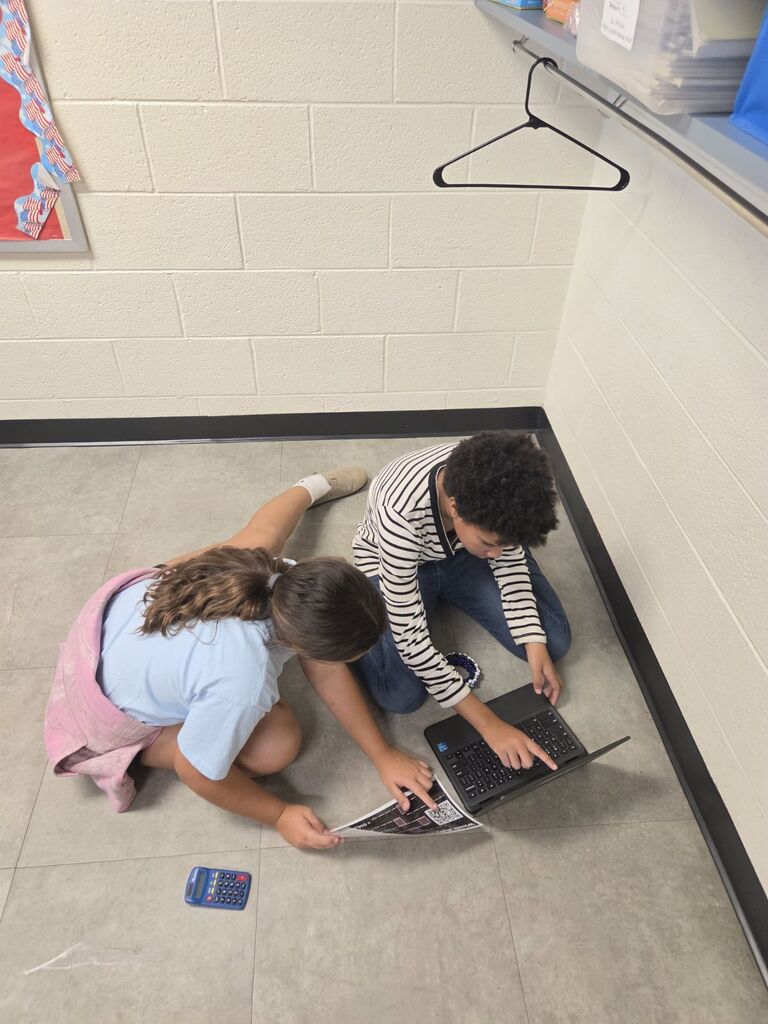 Two individuals seated on the floor, one with a laptop, and a calculator nearby. White walls and a bulletin board.