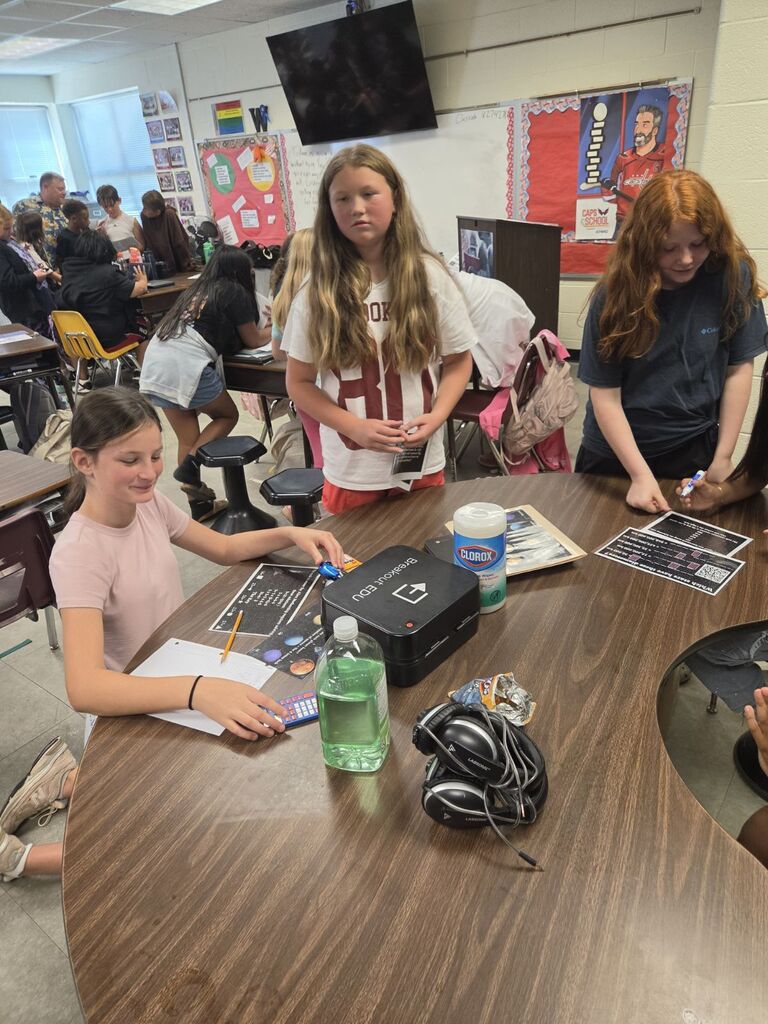 Students gather at a table in a classroom; one sits and writes while two others stand nearby.