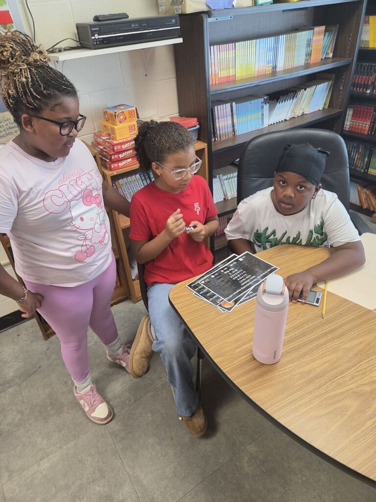 Three children in a classroom, one reading, another looking on, and a third holding a book.
