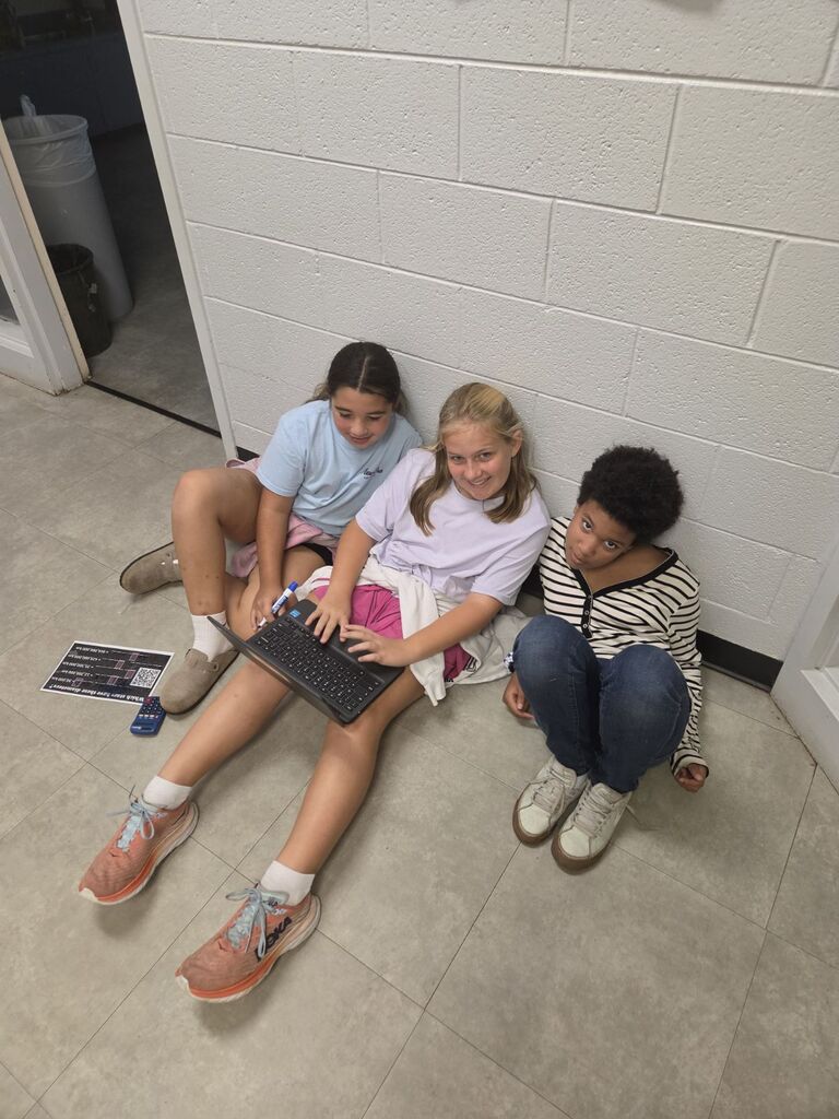Three children seated on floor, one using a laptop, sitting against a white wall with tiled flooring.