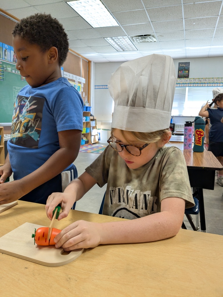 Two children in a classroom, one wearing a chef's hat, chopping a carrot on a cutting board.