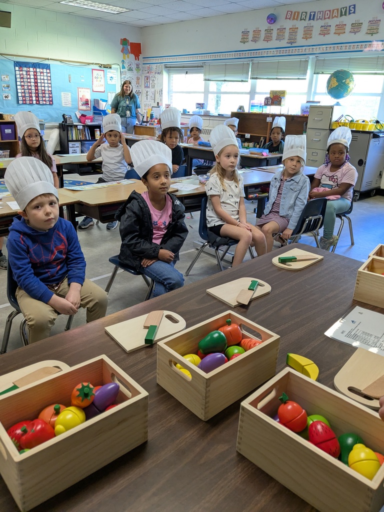 Children in a classroom wearing chef hats, sitting around a table with boxes of colorful toy vegetables.