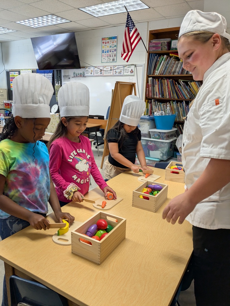 A woman in a chef's hat helps four children in chef hats cut vegetables at a table.