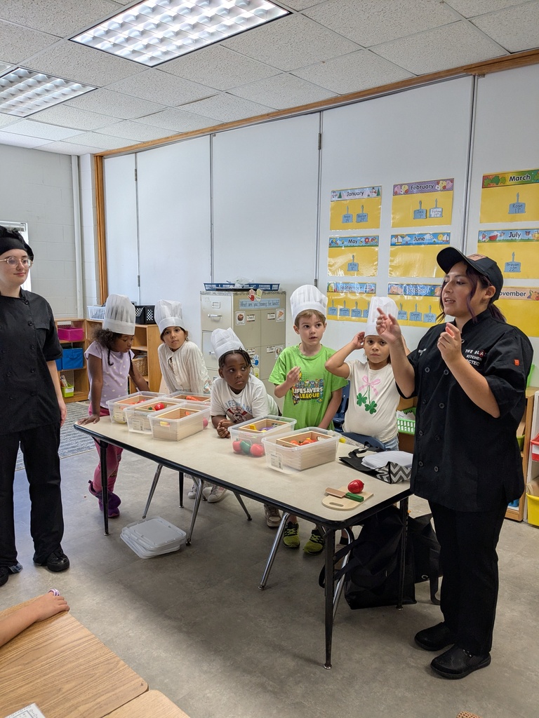 A chef instructs a group of children in chef hats around a table with various food items.