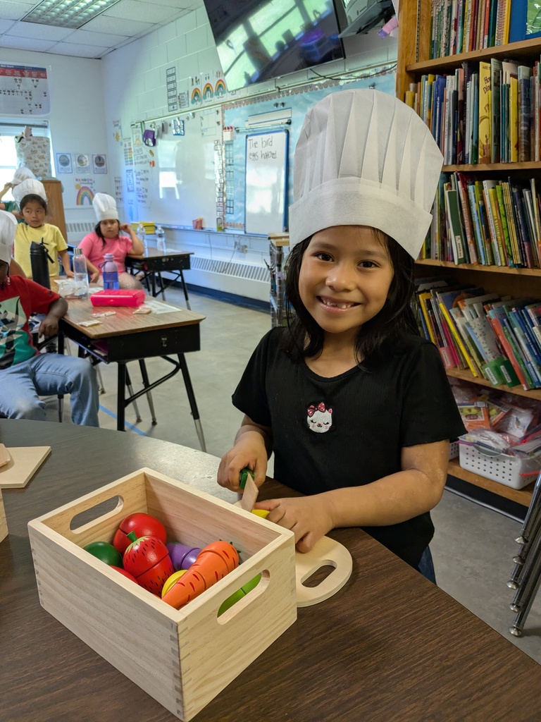A young child wearing a chef's hat sits at a table with a wooden box of vegetables.