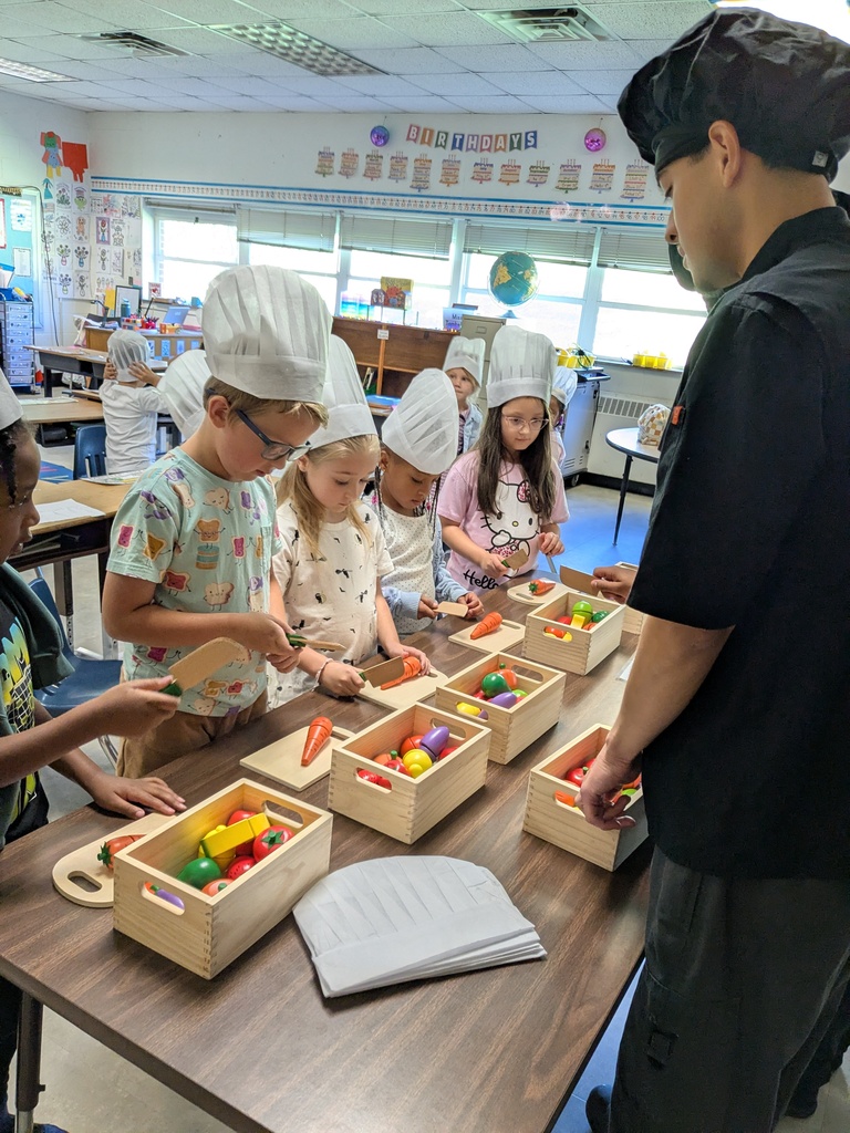 Several kids wearing white hats and aprons are cutting vegetables under the guidance of an adult.