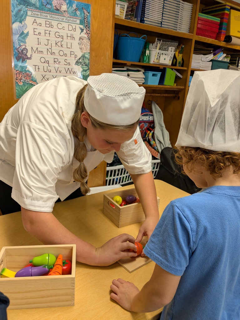 Woman in chef's hat showing a child how to prepare carrots. Background includes a wooden bookshelf.