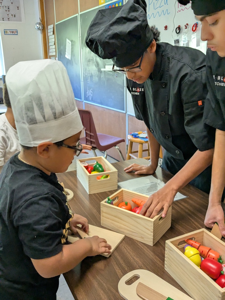 Kids in chef hats cutting vegetables with assistance from adults in a classroom.