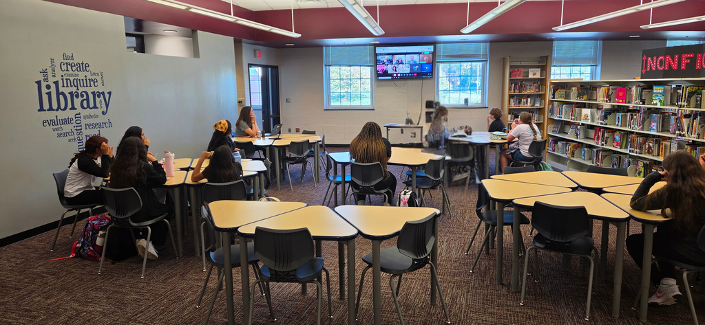 A school library with round tables, chairs, and bookshelves. A television is on the wall.