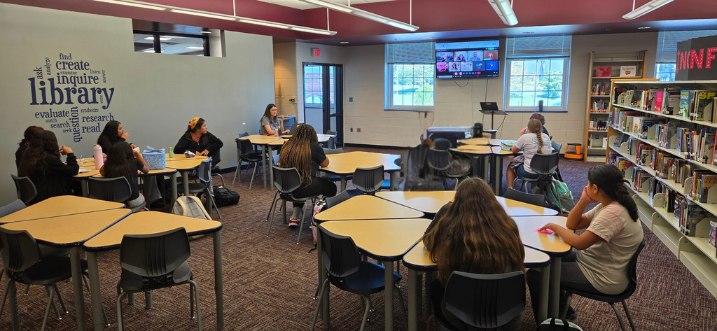 Library with students sitting at tables, a TV, bookshelves, and a beige carpet. White walls with the word "Library" and lamps.