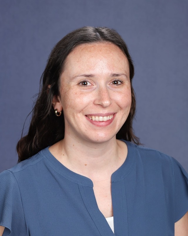 A woman with dark hair, wearing a blue top and earrings, smiles in front of a blue background.