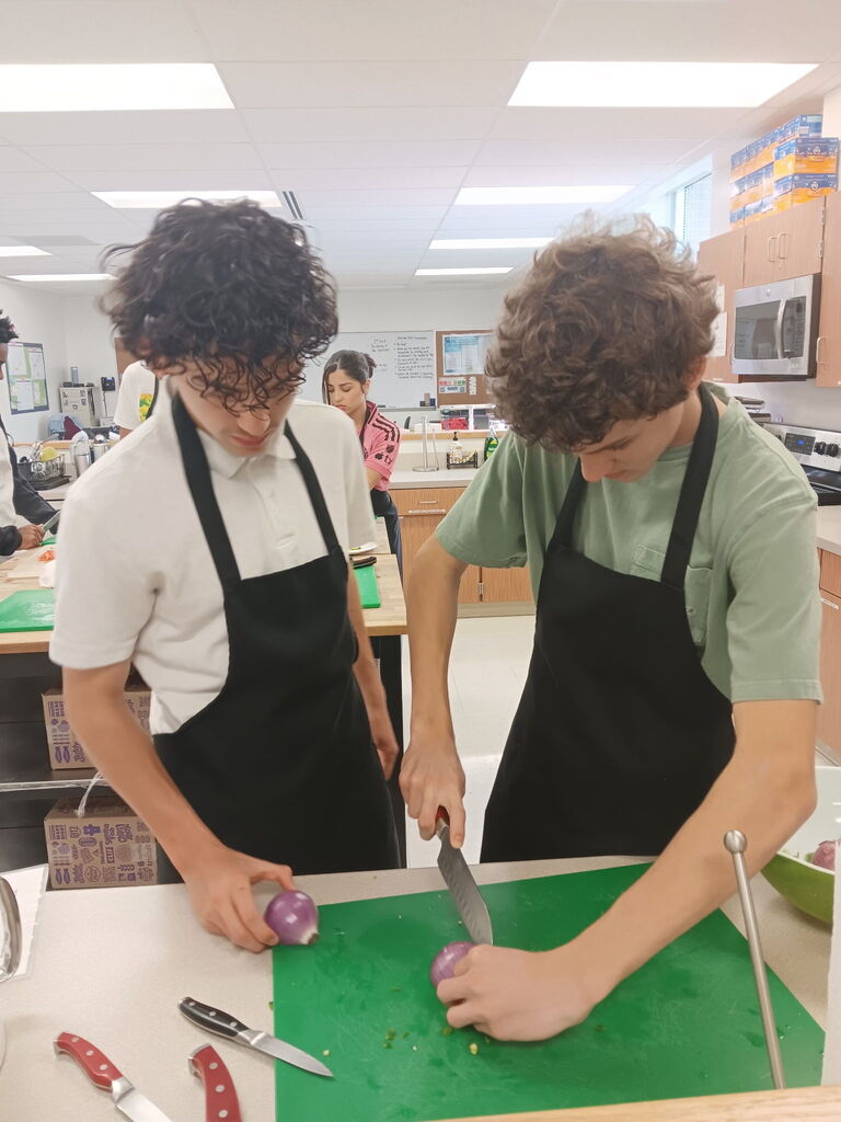 Two individuals are in a kitchen. They wear aprons. One is cutting an onion on a green board.