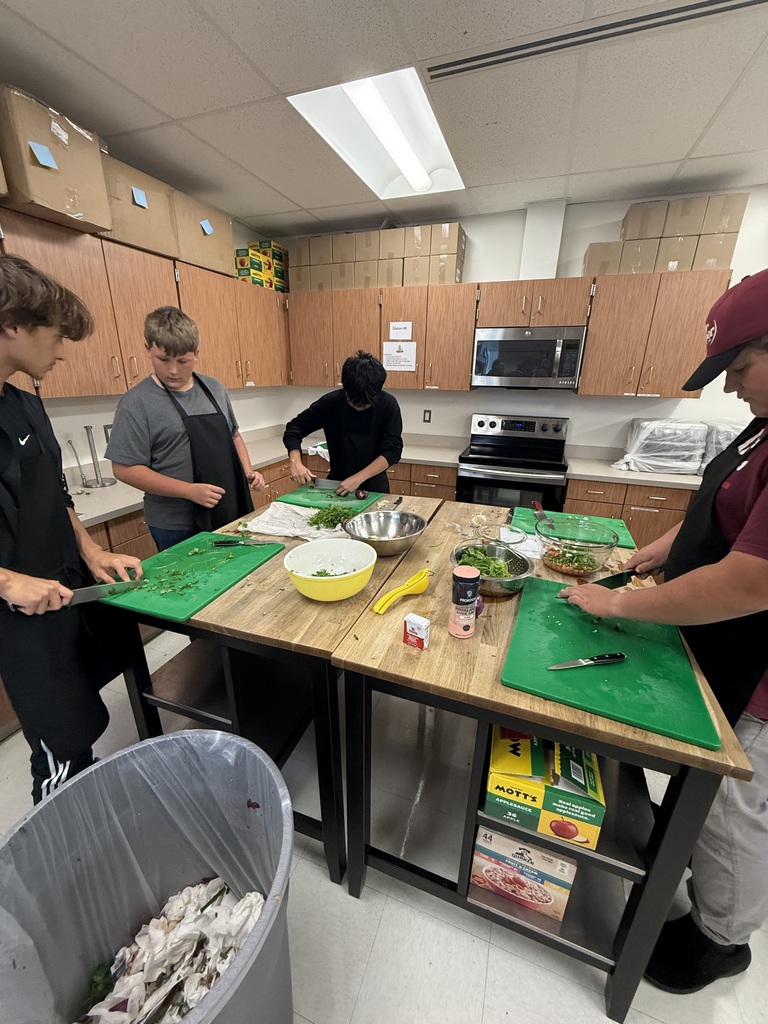 Four people in aprons prepare food at a table in a kitchen with wooden cabinets.