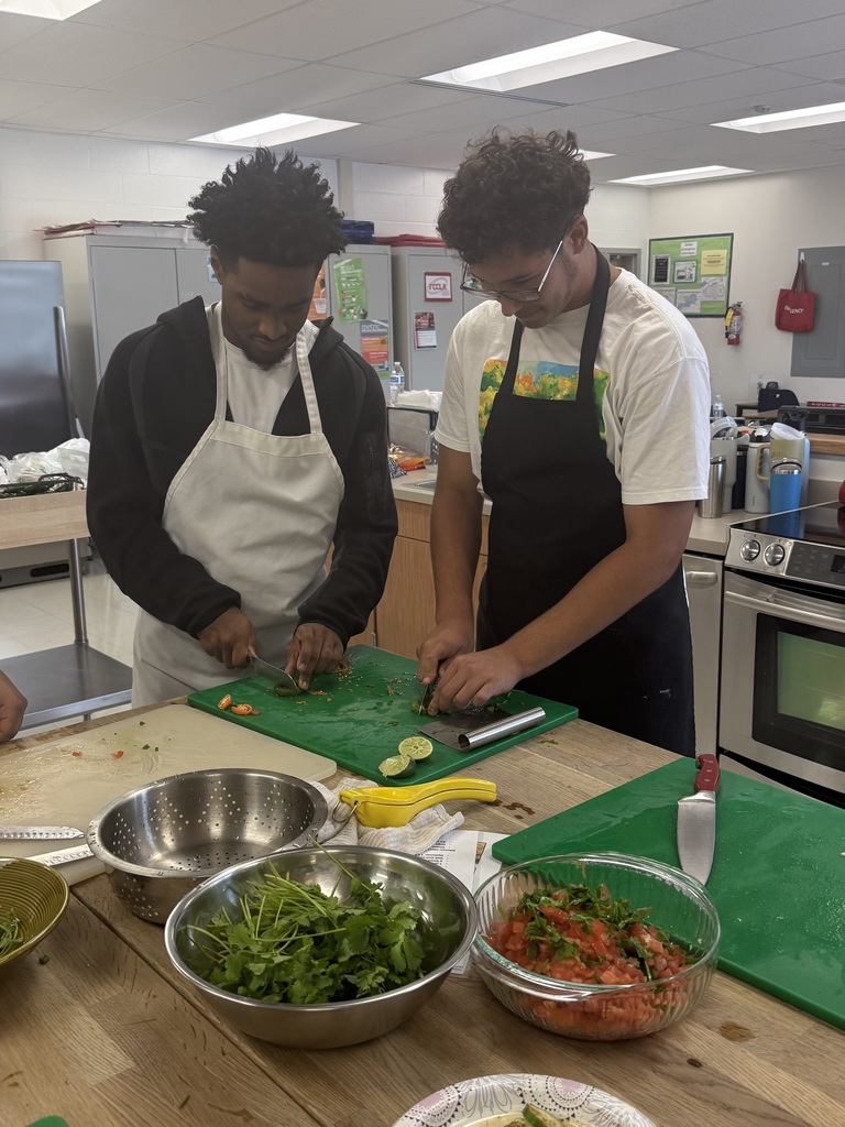 Two people in aprons are cutting food on a table with bowls of ingredients.