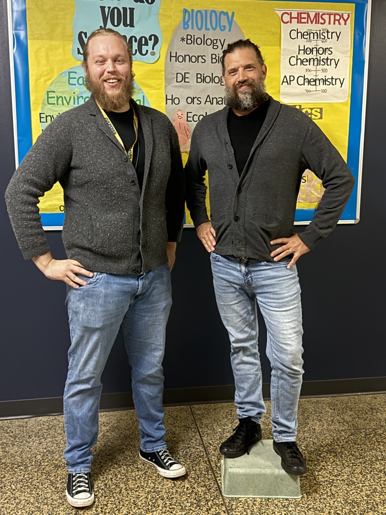 Two men pose in front of a bulletin board with text that reads "Biology" and "Chemistry" in a classroom.