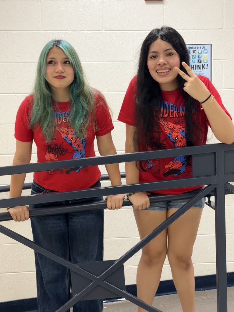 Two women with different hair colors, one with green, standing behind a railing. Both wear red shirts with a Spider-Man graphic.